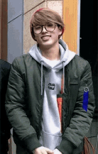 a young man wearing glasses and a green jacket is standing in front of a building and smiling .