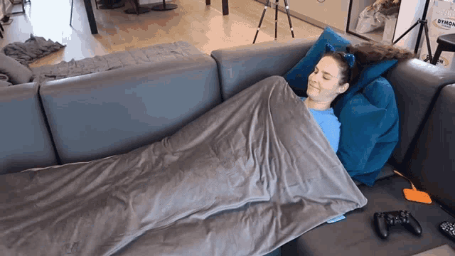 a woman wrapped in a blanket is laying on a couch with a game controller in front of her