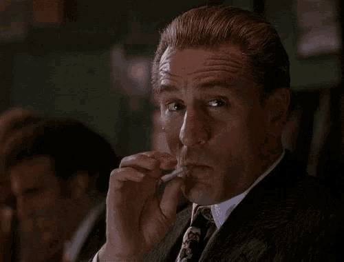 a man in a suit smoking a cigarette with another man in the background