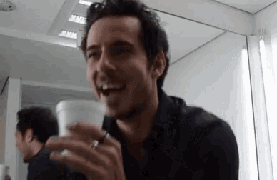 a man in a black shirt is holding a cup of coffee and smiling in front of a mirror .