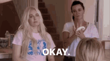 two women are standing next to each other in a room and one of them is holding a napkin and says `` okay '' .