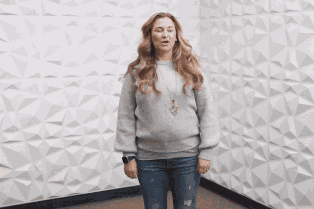 a woman wearing a grey sweater and jeans is standing in front of a white wall