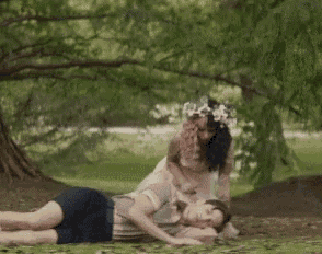 a man and a woman are laying on the grass in a park . the woman is wearing a flower crown .