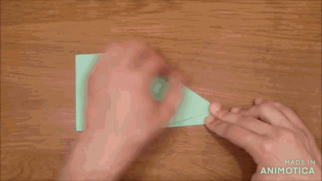 a person is folding a piece of green paper on a wooden table and the words made in animatica are visible