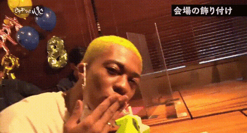 a man with yellow hair is sitting at a table with balloons in the background and eating a banana .