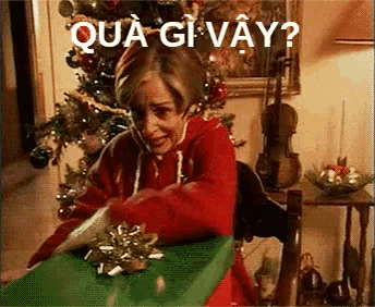 a man in a red sweater is sitting in front of a christmas tree opening a gift .