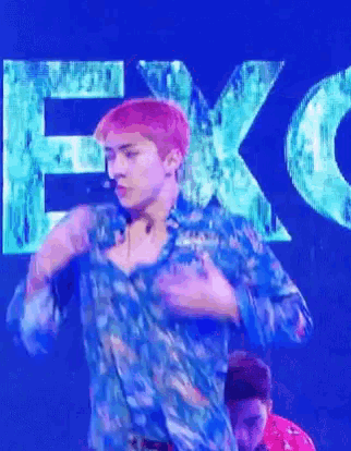a man with pink hair is wearing a blue shirt and dancing on a stage in front of a sign that says exo .
