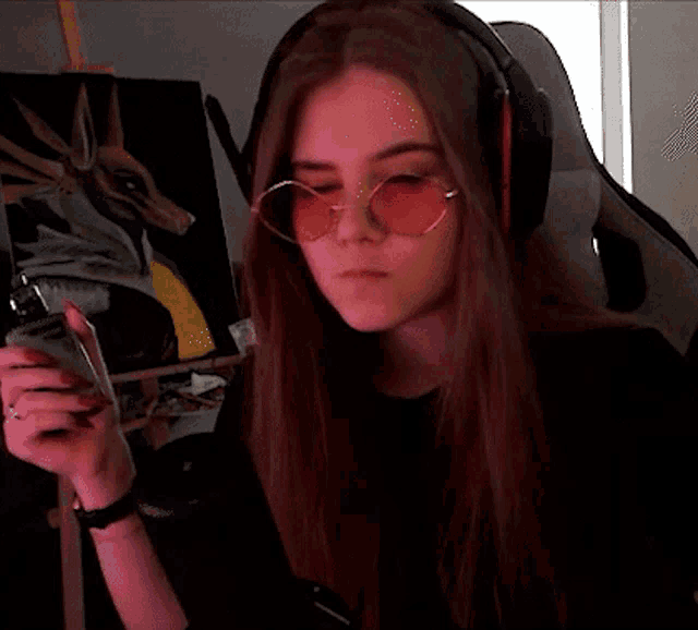 a girl wearing headphones and sunglasses is sitting in front of a painting and holding a cell phone .