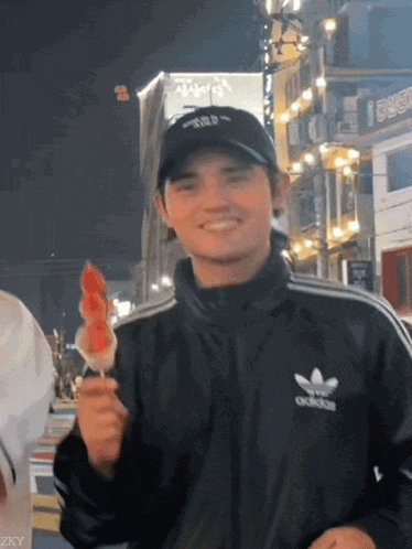 a man wearing a black adidas jacket holds a watermelon on a stick