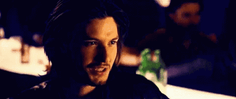 a man with long hair and a beard is sitting at a table in a dark room and looking at the camera .