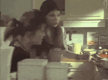 two women are standing in a kitchen talking to each other while a can of corn is in the background .