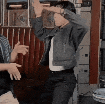 a woman in a denim jacket is dancing in a diner with her hands in the air