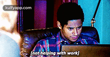 a man in a plaid shirt is sitting in front of a laptop computer and says " not helping with work "