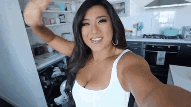 a woman in a white tank top is taking a selfie in a kitchen .