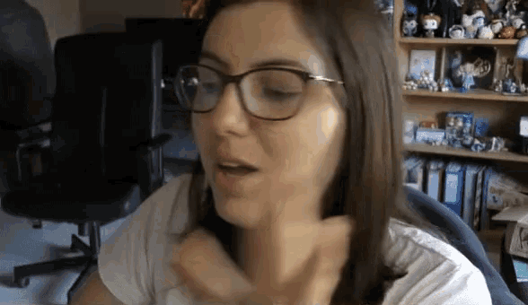 a woman wearing glasses is sitting at a desk in front of a bookshelf filled with figurines .
