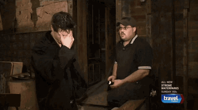 two men are standing in a dark room with a travel channel ad behind them