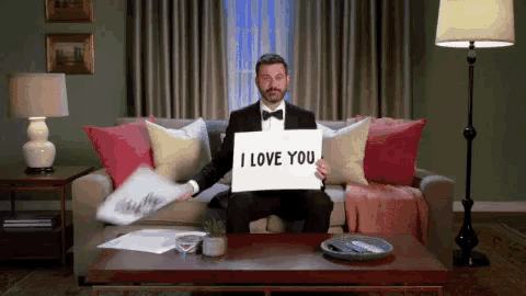a man in a tuxedo is sitting on a couch holding a sign that says " i love you "