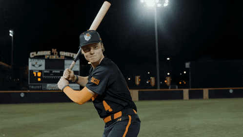 a baseball player holding a bat in front of a scoreboard that says claude smith field