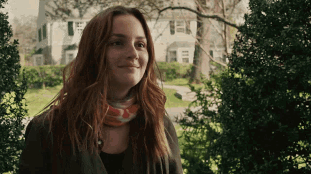 a woman with red hair and a scarf around her neck smiles in front of a white house