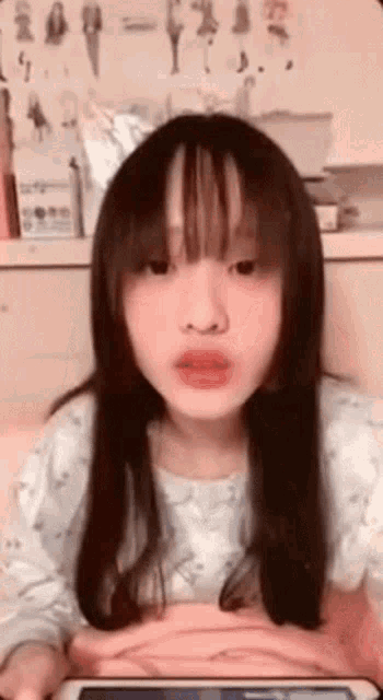 a young girl with long hair and bangs is looking at the camera while holding a tablet .