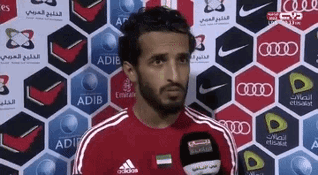 a man wearing a red adidas shirt stands in front of a wall of advertisements including emirates and audi