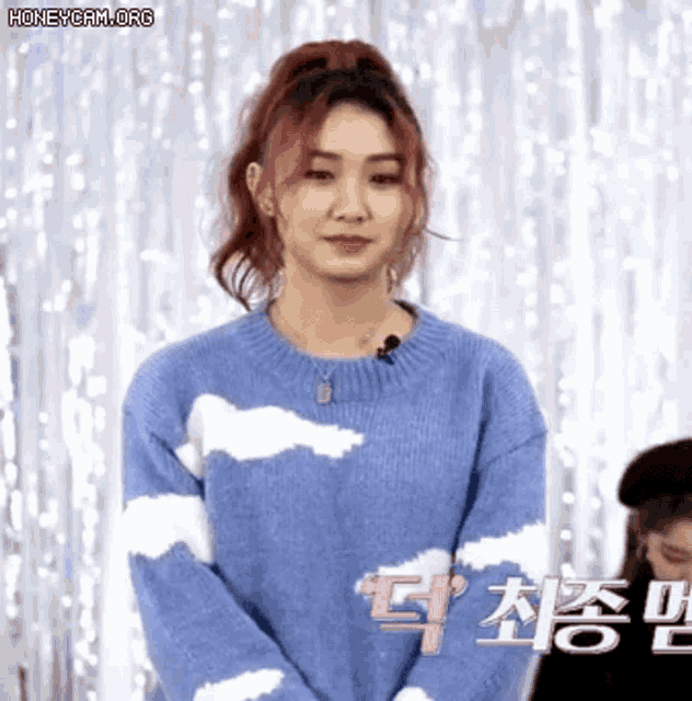 a woman wearing a blue sweater with white clouds on it is standing in front of a white curtain .