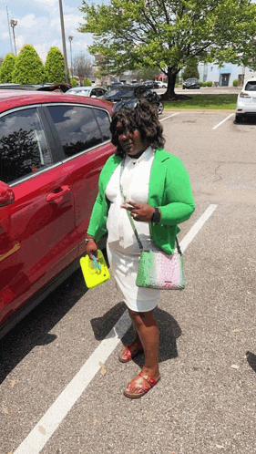 a woman in a green jacket and white dress stands in a parking lot next to a red car