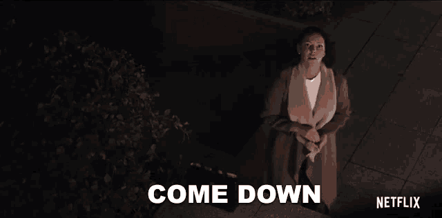 a woman in a coat is standing on a sidewalk at night and says come down .