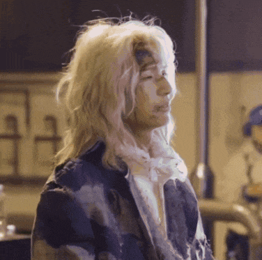 a woman with long blonde hair and white paint on her face is wearing a dirty jacket .