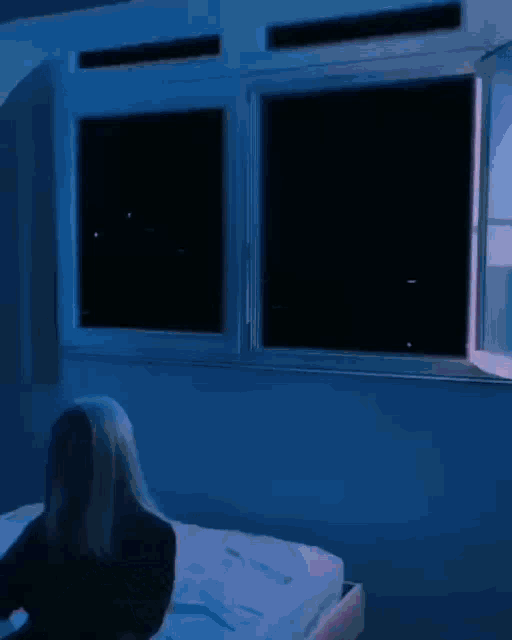 a woman is laying on a bed in a dark room with two windows open