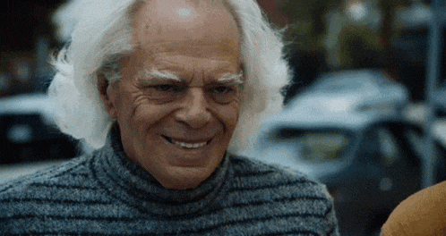 an elderly man with white hair is smiling and wearing a gray sweater .