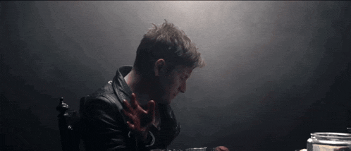 a man in a leather jacket sits at a table with his hands in the air