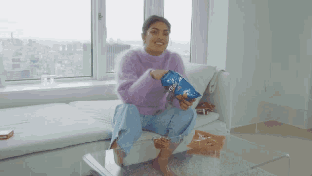 a woman in a purple sweater is sitting on a couch holding a bag of doritos chips