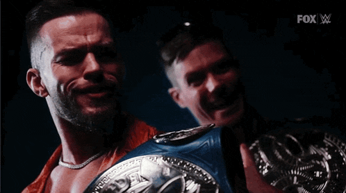 two men are standing next to each other and one of them is holding a championship belt .