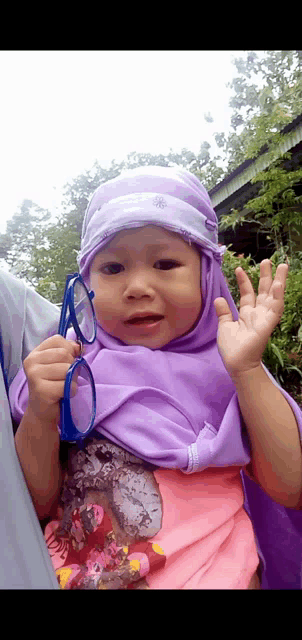 a little girl wearing a purple hijab holds a pair of sunglasses