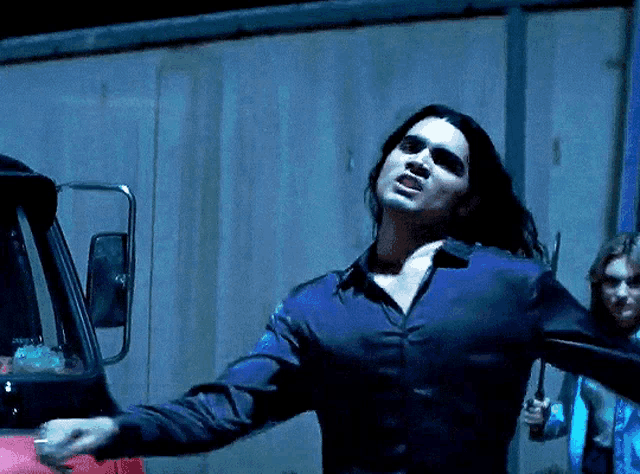 a man with long hair in a blue shirt stands in front of a truck