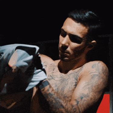 a shirtless man with tattoos on his arms holds a shirt up