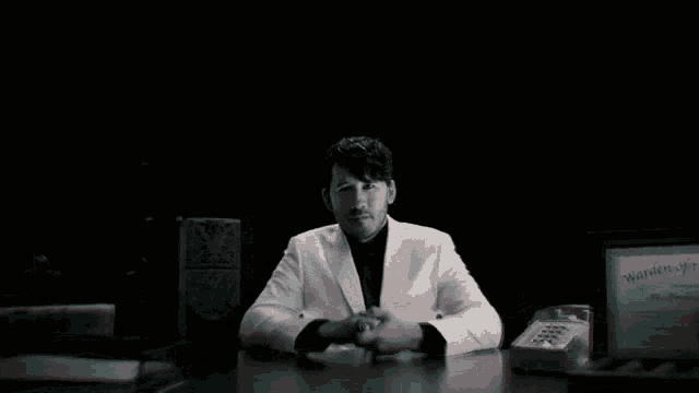 a man in a white suit sits at a desk in front of a sign that says ' witness of the will '