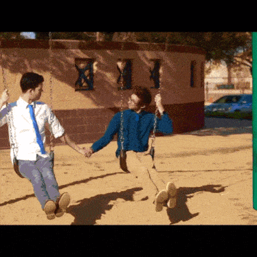 two men are holding hands while sitting on swings in the sand