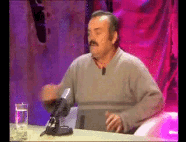 a man with a mustache is sitting at a table with a microphone in front of him .