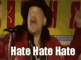 a man in a hat is standing in front of a microphone with the words `` hate hate hate '' written above him .