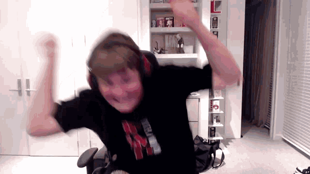a man wearing headphones and a black shirt with the letter k on it is sitting in a chair with his arms in the air