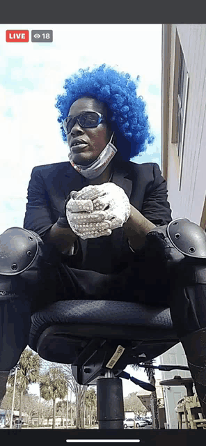 a man wearing a blue wig and gloves is sitting in a chair with a live button on the bottom