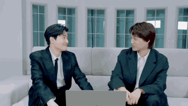two men in suits and ties are sitting on a couch looking at a laptop