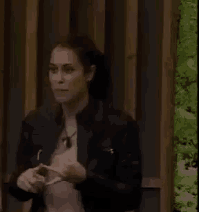 a woman in a black jacket is standing in front of a wooden fence holding a cell phone .