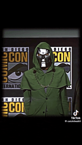 a man in a green hooded jacket stands in front of a comic con poster