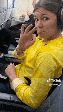 a woman wearing headphones and a yellow shirt is sitting on an airplane and making a peace sign