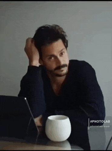 a man with a mustache is sitting at a table with a cup of coffee and a laptop