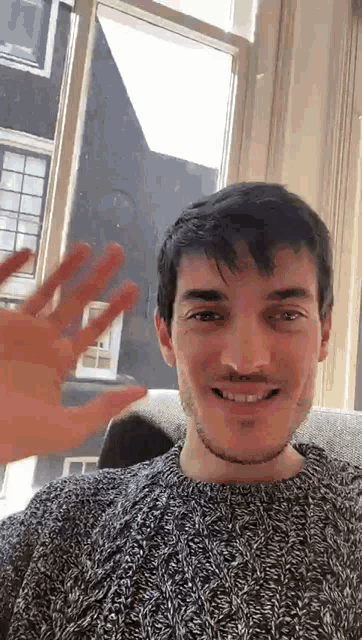 a man in a grey sweater is smiling and waving his hand