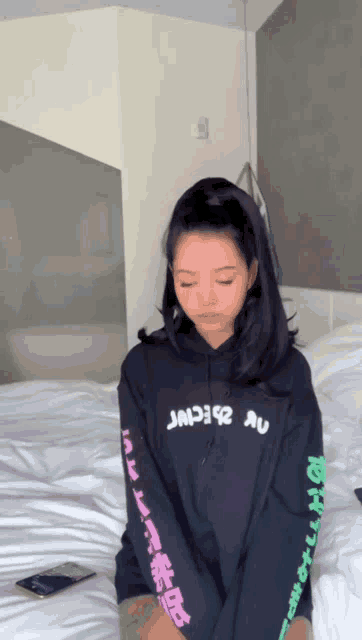 a girl wearing a black hoodie with chinese writing on the sleeves is sitting on a bed